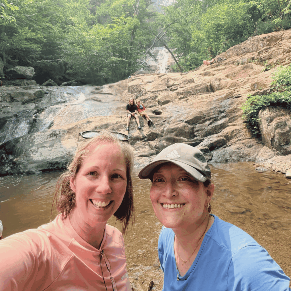 Marcy Holbert and her best friend smiling beside a waterfall along Whiteoak Canyon Trail in Shenandoah National Park, with their sons exploring the rocks behind them.